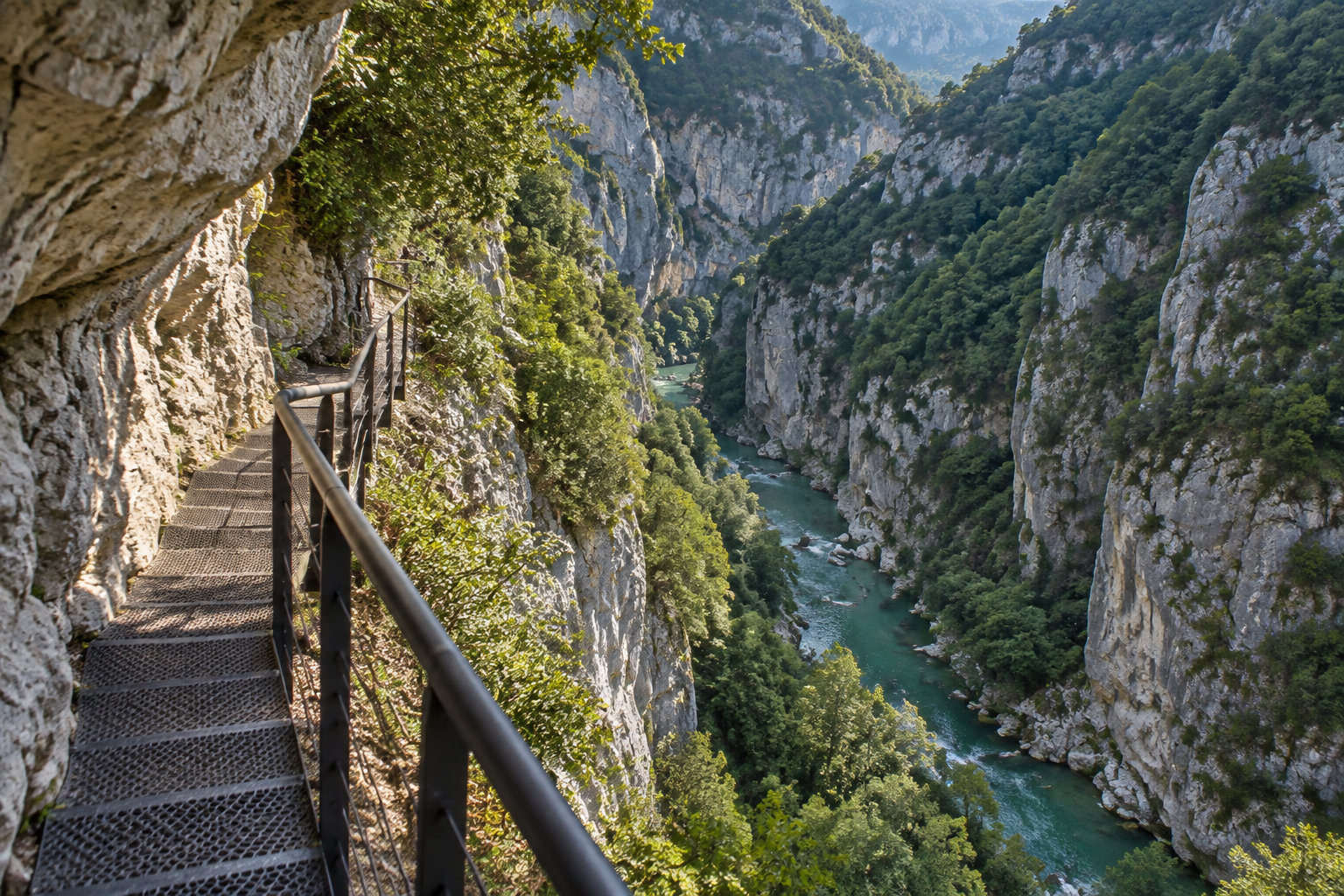 Un escalier métallique à flanc de falaise surplombe une rivière turquoise encaissée dans des gorges rocheuses et boisées.