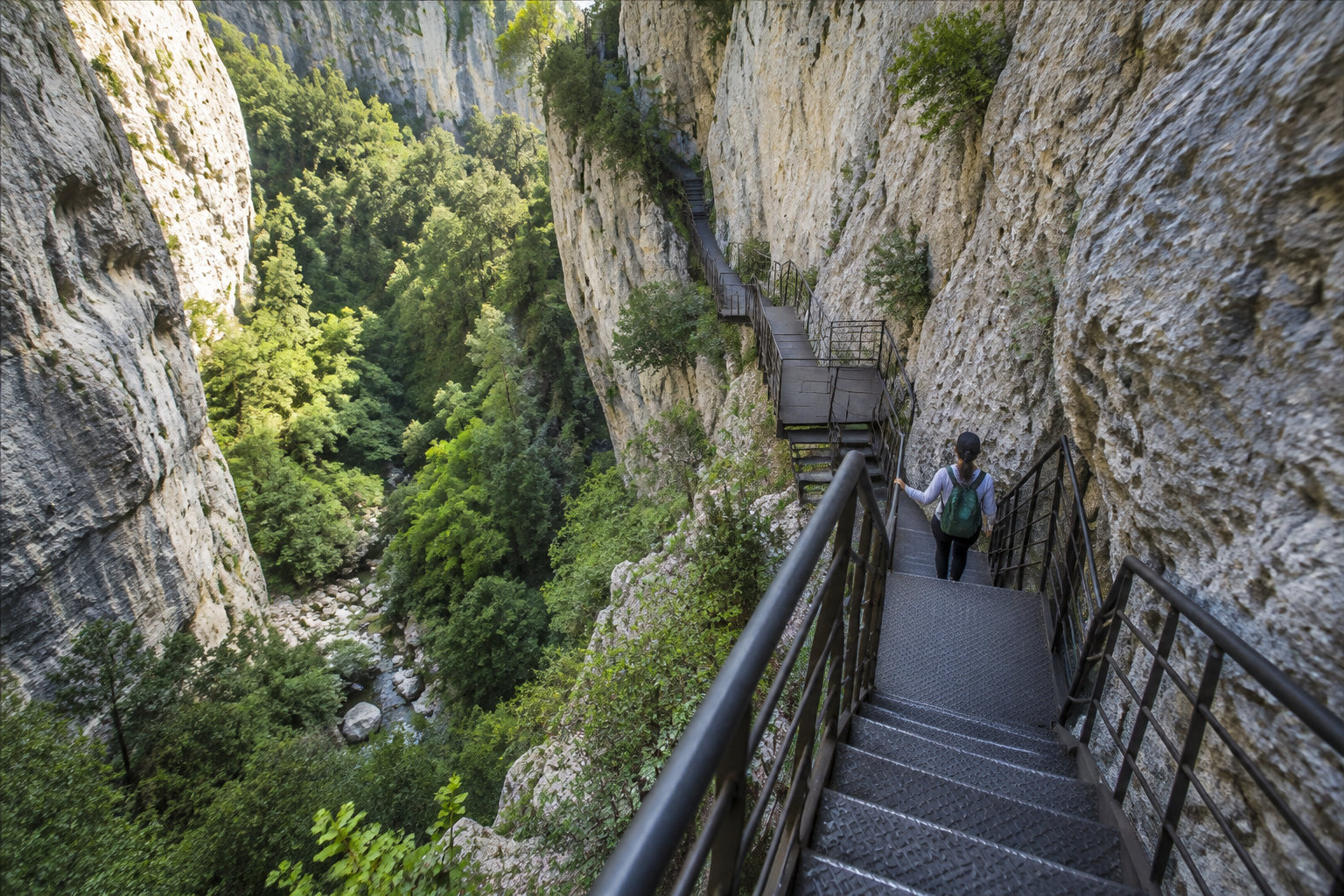 Une personne portant un sac à dos descend un escalier métallique fixé à une falaise, au-dessus d’une gorge boisée.