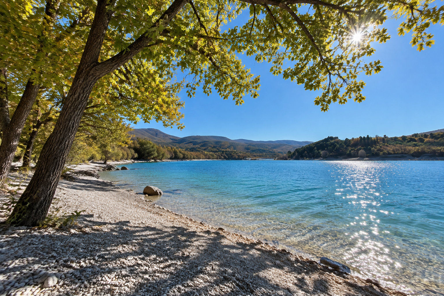 Un lac aux eaux bleues scintille au soleil, bordé d’une rive rocheuse et d’arbres feuillus, avec des montagnes à l’horizon.