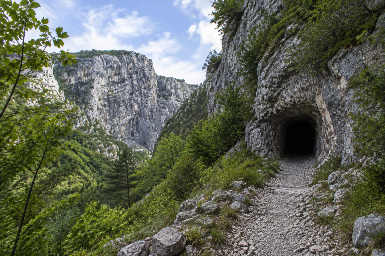 Un sentier rocailleux mène à l’entrée sombre d’une grotte dans une falaise, au milieu d’une végétation verdoyante et de montagnes rocheuses.