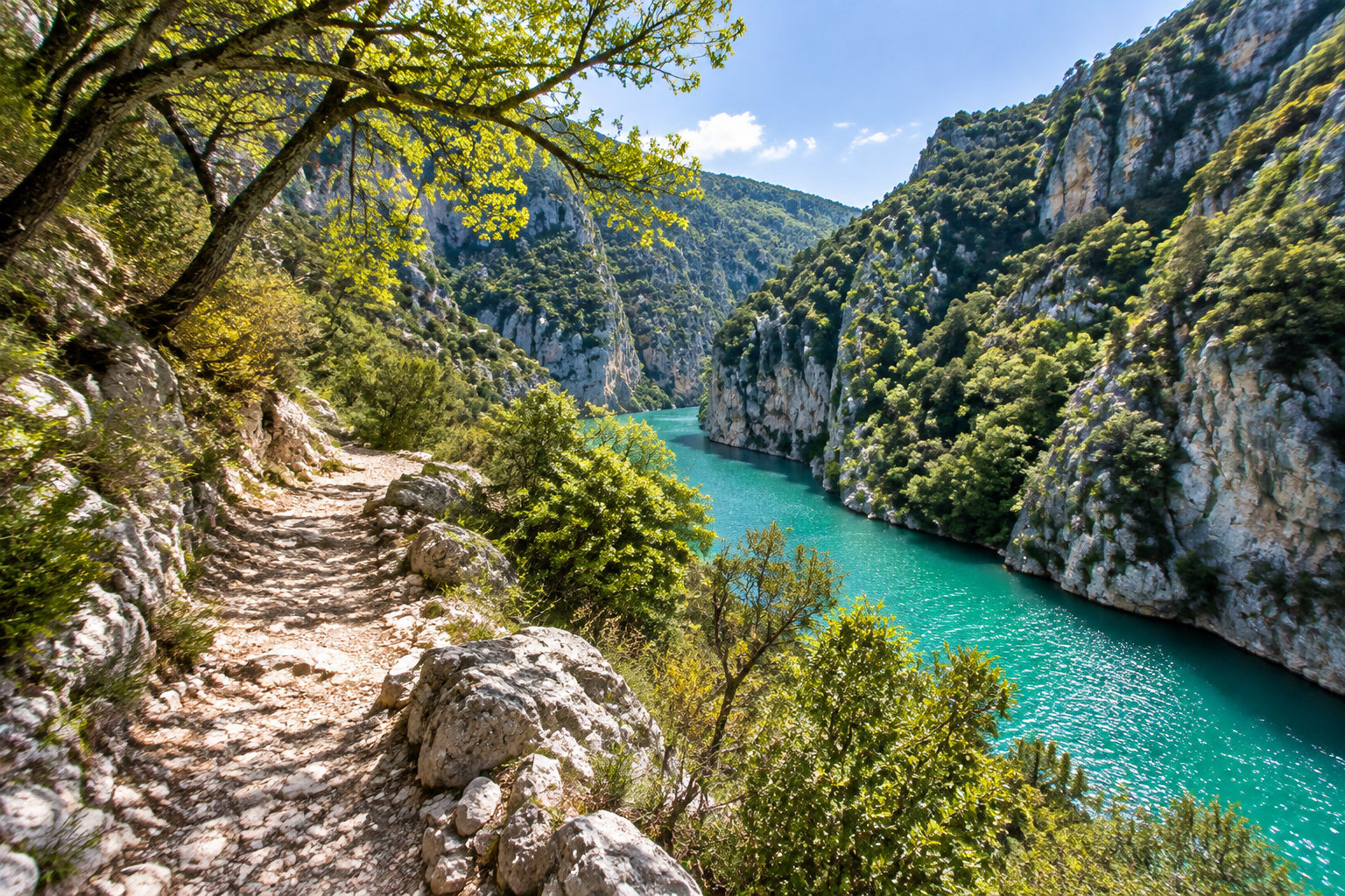 Un sentier rocailleux longe une rivière turquoise au fond d’une gorge bordée de falaises boisées.